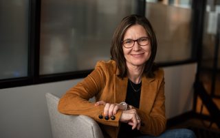 Lisa Carmichael wearing a jacket and her glasses with her arm on a sofa